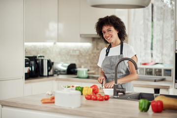 Gorgeous mixed race housewife in apron standing in kitchen and chopping mushrooms. On kitchen counter are all sorts of vegetables.