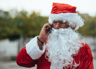 Stock photo of Santa claus talking to a mobile phone with a park behind unfocused. Christmas time