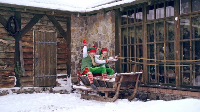 Christmas elves ride wooden sledges. New Year is coming. Christmas eve. Boy and girl wear christmas clothes. Having fun.