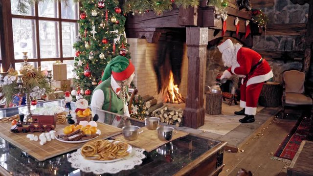 Santa Claus and an elve girl. Santa preparing tea. Santa Claus with boiling teapot, Curious girl wears elves clothes. Happy elf girl in fairy costume sitting near the table. Santa near Christmas firep