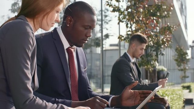 Medium shot of young businesspeople of different ethnicities sitting on bench and discussing something they see on laptop computer screen, man in the background using smartphone
