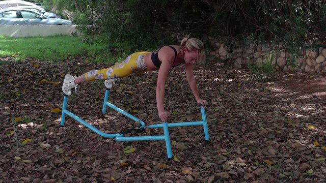 Athletic woman with a muscular body and blonde hair doing push-ups, using parallel bars. Urban park background.