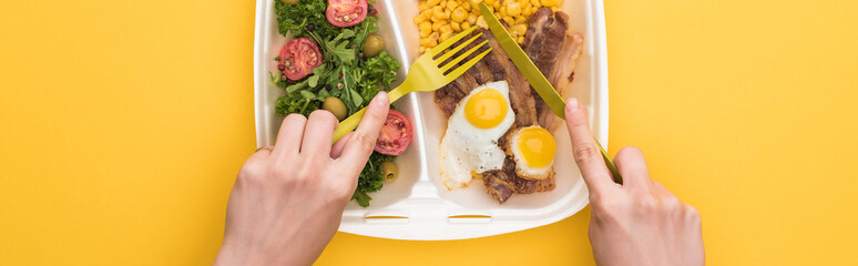 panoramic shot of woman eating from eco package with corn, meat, fried eggs and salad isolated on yellow