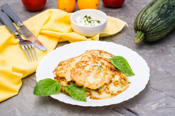 Vegetable fried fritters from zucchini and basil leaves on a plate and sour cream sauce with greens in a bowl on the table