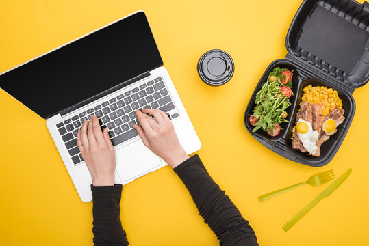 cropped view of woman using laptop and eco package with corn, meat, fried eggs and salad, paper cup isolated on yellow