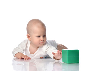 Curious child baby toddler in white bodysuit is lying on his stomach, touching green cube, playing, pushing it on white