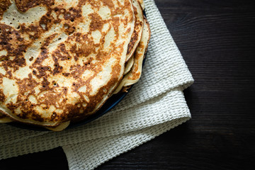 A stack of baked pancakes in a plate on a folded kitchen towel is white in gray. Dark wooden surface. Horizontal orientation.