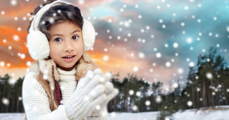 christmas, season and people concept - happy little girl wearing earmuffs over winter forest background