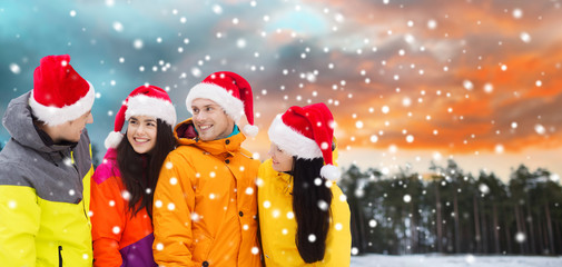 holidays, christmas, friendship and people concept - happy friends in santa hats and ski suits over winter forest background