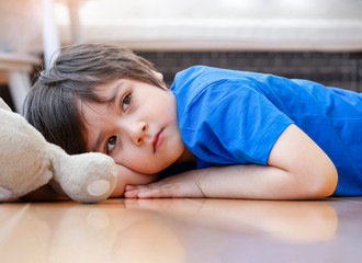 Portrait of Lonely kid with sad face lying down on wooden floor with his teddy bear in playroom at...
