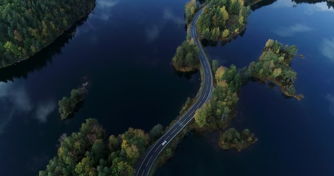Drone follows white car wich drives the road between lakes. Sky and clouds are reflected in the water.