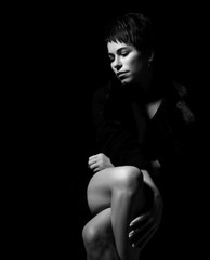 Black and white portrait of sad woman feeling grief sitting in darkness with her legs crossed looking down on dark