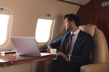 pensive businessman holding cup of coffee in plane with laptop during business trip