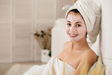 smiling woman with towel is applying mask on her face and looking at the camera at home. Skin care. space for text