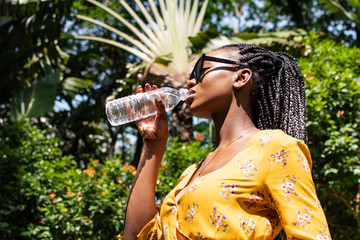 Woman drinking water from plastic water bottle. Woman wearing yellow dress. Palm tree, sunglasses, side on view. Beautiful African woman. Clear bottle.