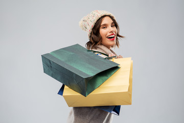 christmas, seasonal sale and consumerism concept - happy smiling young woman in knitted winter hat and sweater with shopping bags over grey background