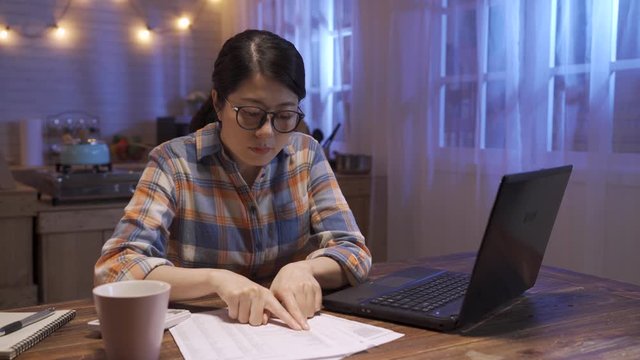 beautiful asian korean business lady work day and night. woman in casual wear sitting at dark home kitchen type on laptop computer and counting on calculator with document papers. female hardworking
