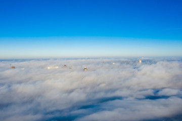 Obraz premium Quadrocopter flying over the fog at high altitude over the city. The upper floors of high-rise buildings among huge clubs of white fog with a horizon line on a crystal clear sky.