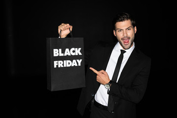 excited man showing shopping bag with black friday sign, isolated on black