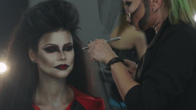 blonde woman artist in image makes halloween makeup to model using airbrush preparing for party close view