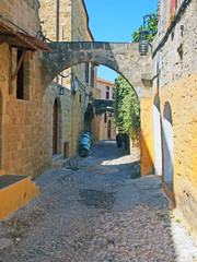 typical quiet cobbled street in rhodes town with old yellow painted houses and a stone arch with sunlit summer sky