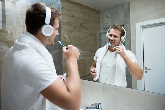 young handsome smiling man in headphones brushing teeth in bathroom