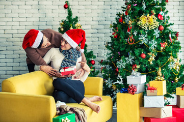 Happy young adult man hugging beautiful woman girlfriend and surprise her a Christmas present in gift box with Christmas tree decoration background. Christmas family celebration festive party concept.