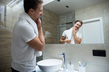 young handsome smiling man with white towel on his shoulders looking in the bathroom mirror