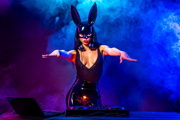 Young sexy woman dj playing music in mask. Headphones and dj mixer on table. Smoke on background