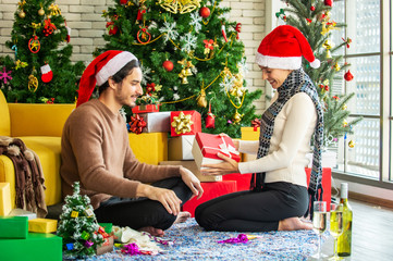 Obraz na płótnie Canvas Happy young adult man in santa hat give a Christmas present in gift box to beautiful woman girlfriend with Christmas tree decoration background. Christmas family celebration festive party concept.