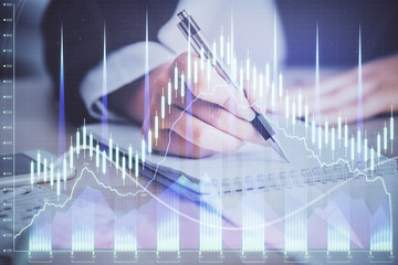 A woman hands writing information about stock market in notepad. Forex chart holograms in front. Concept of research. Multi exposure