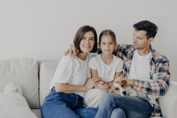 Family time concept. Cheerful mum, dad and small girl embrace all together, spend spare time in living room of their new home, sit on sofa, play with dog, have perfect relationships, enjoy comfort