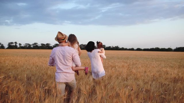 happy family couple in white runs along boundless wheat field holding small daughters in arms under blue sky slow motion