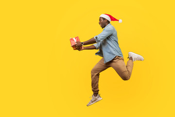 Full length side view of joyous handsome young man in santa hat and casual denim shirt jumping or running with christmas gift box, hurry for sales. indoor studio shot isolated on yellow background