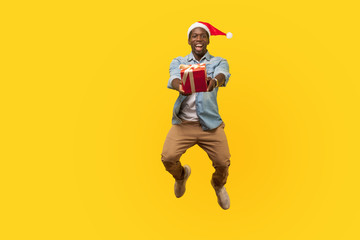 Fototapeta na wymiar Full length portrait of extremely happy young man in santa hat and casual denim shirt jumping or flying and giving christmas gift box to camera. indoor studio shot isolated on yellow background