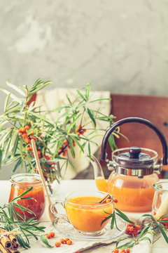 Cup and teapot of hot spicy tea with sea buckthorn, jam in the glass jar, branches of fresh berries on light wooden table surface in the rustic room