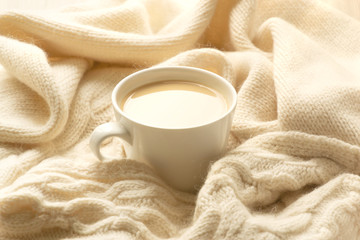 The white cup of hot coffee or tea with milk and the ivory color warm plaid. Comfort, cosiness and warmth concept. Picture in light pastel colors