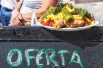 Food to eat at the street. Delicious dish over a sign with the text "Oferta" (offer in spanish language). Menu with chorizo, green peppers, chips and ribs. Sunny scene of warm tone.