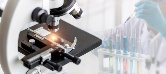Close-up shot of microscope with metal lens at laboratory with blurred background of scientist use dropper to transfer chemical reagent to test tube. He observe the chemical reaction in laboratory.