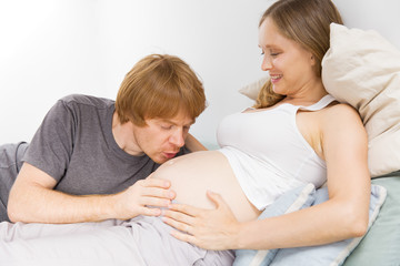 Cheerful expectant couple feeling happy at home. Young man talking to pregnant wife belly. Baby talk concept