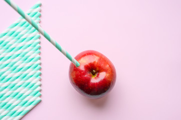 a ripe juicy bright red apple and in it a striped white and green paper drinking straw, and a pile of drinking straws nearby, located on a pink background