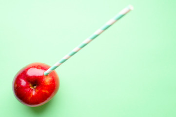 a ripe juicy bright red apple and in it a striped white and green paper drinking straw, located on a green background. space for text