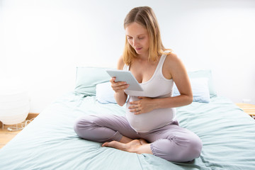 Positive expectant mom consulting internet on tablet. Pregnant woman sitting on bed, using digital device, looking at screen. Communication or social network concept