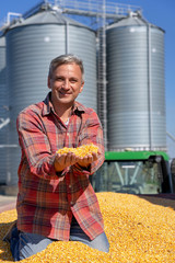 Farmer Sitting in Trailer Full of Freshly Harvested Corn Seeds Against Grain Silo