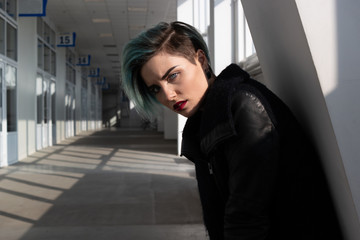 Obraz premium Portrait of beautiful girl with short blue fashionable hairstyle and red lipstick in black coat in a long white corridor with many doors and windows.