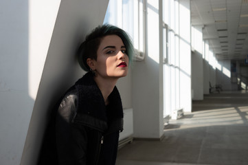 Beautiful girl with short blue fashionable hairstyle in black coat leans against the column in a long white corridor. Model looking at the camera and shadow hiding part of her face.