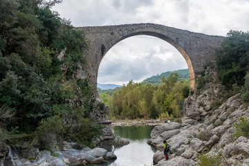 Fototapeta na wymiar A young hiker contemplates the old medieval bridge of Llierca, in the region of La Garrotxa, Girona, Spain, on a cloudy dramatic day.