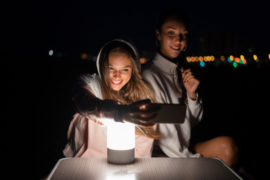 Two female, friends with led light on camping table at night taking a selfie