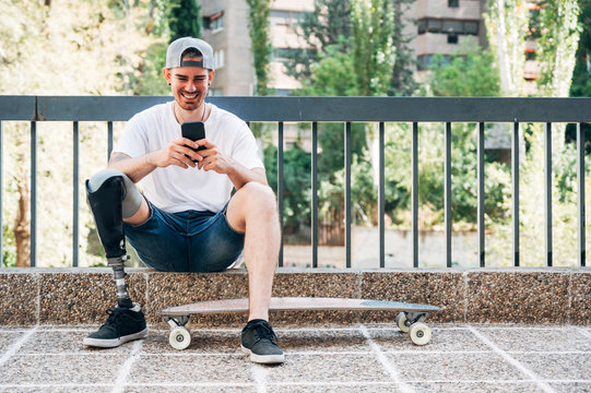 Smiling young man with leg prosthesis and skateboard using smartphone