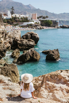 Back view of woman wearing white dress and straw hat sitting on rocks looking to the sea, Nerja, Spain